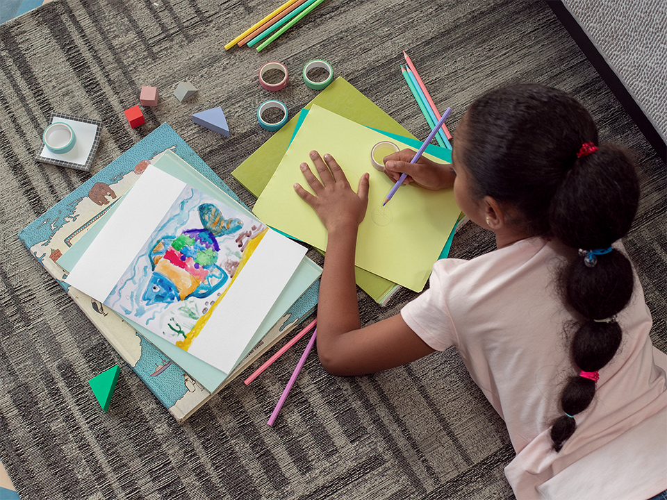 A child drawing on a yellow paper with crayons and geometric shapes scattered around.